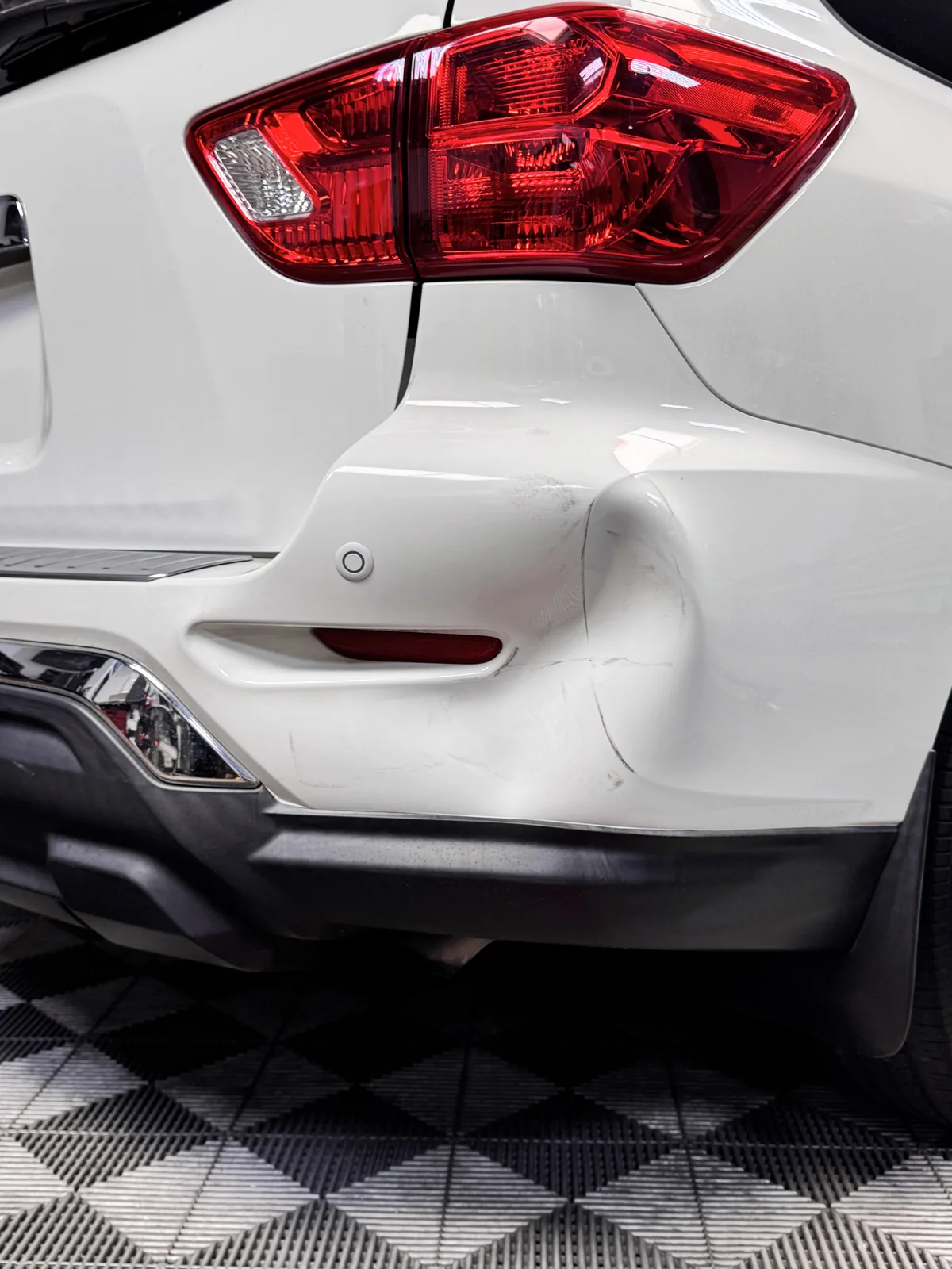 A deep dent in the rear bumper corner of a white Nissan SUV with the paint still intact